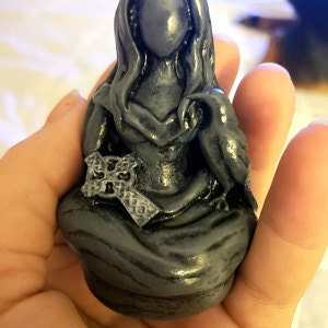 May include: A dark gray figurine of a seated figure holding a cross and a raven. The figure has long hair and is wearing a draped garment. The figurine is held in a person's hand.