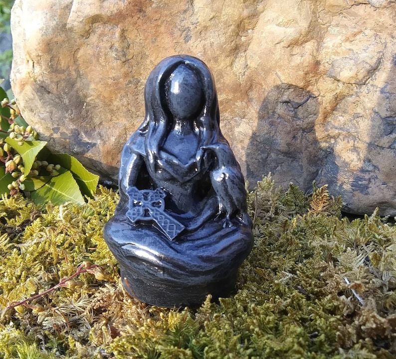 May include: A dark gray statue of a seated figure with long hair, holding a cross and a bird. The statue is set against a backdrop of moss and a large rock, with sunlight illuminating the details of the sculpture.