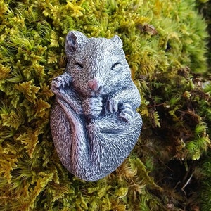 May include: A gray, detailed sculpture of a curled-up squirrel with its eyes closed, resting on a bed of green and yellow moss. The squirrel has a textured surface and a pink nose. The moss provides a natural, earthy backdrop.