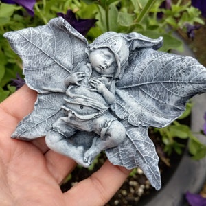 May include: A gray resin sculpture of a sleeping fairy with wings made of leaves. The fairy is lying on its back with its arms crossed over its chest. The sculpture is detailed with realistic features and textures.