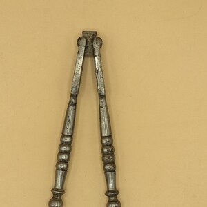 May include: A vintage-style metal nutcracker featuring two long, slender handles with a series of rounded, ribbed segments and spherical ends. The tool has a weathered, dark grey metallic finish with visible signs of wear and oxidation throughout.