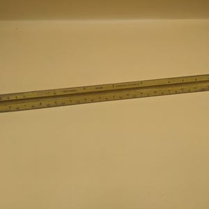 May include: A beige triangular architect scale ruler featuring black numerical markings and graduation lines. The ruler is inscribed with the text KEUFFEL & ESSER CO. 2428 ENGINE DIVIDED. It has a three-sided profile with distinct edges for measurements.