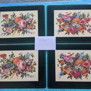May include: A set of four rectangular placemats featuring a dark green border with a thin gold line accent. The center of each mat displays a cream-colored panel with a detailed floral and fruit arrangement including pink roses and oranges.