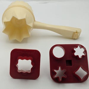 May include: A vintage Tupperware cookie press with a cream-colored handle and flower-shaped press. Two red cookie cutters with various shapes are also included, perfect for baking. The Tupperware logo is visible on one of the cutters.