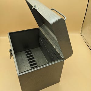 May include: A gray metal storage box with a hinged lid and a top handle. The interior features a series of vertical slots along the sides and base, designed for organizing items. The box includes a metal latch on the front to secure the lid closed.