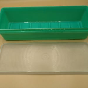 May include: A rectangular, teal-colored plastic container with a matching lid. The container has a ridged interior and a slightly raised rim. The lid is a light gray color and sits next to the container.