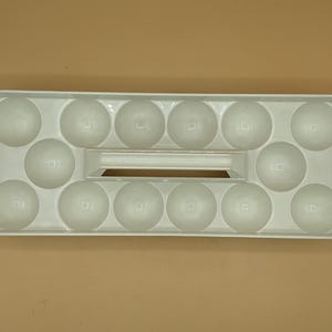 May include: A white plastic egg holder with space for 14 eggs. The rectangular tray has a handle in the center and individual rounded compartments for each egg. The egg tray is on a tan surface.