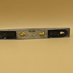 May include: A silver-colored level tool with two yellow bubble vials and a green vial. The level has black end caps and a keyhole-shaped opening on one end. The tool is designed for measuring horizontal and vertical alignment.