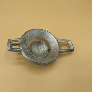 May include: A silver-colored metal object with a circular design and two rectangular extensions on either side. The center features a smaller, rounded shape within the larger circle. The object appears to be a decorative or functional piece.