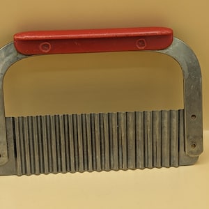 May include: A kitchen crinkle cutter featuring a rectangular metal blade with a wavy, serrated edge. The blade is attached to a U-shaped metal frame with a smooth, solid red handle fixed across the top for a secure grip during food preparation.