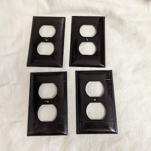 May include: Four rectangular black electrical outlet wall plates, each featuring two circular openings for a standard duplex receptacle and a single centered screw hole for mounting, arranged in a two-by-two grid formation.