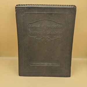 May include: A dark gray, rectangular instruction manual for a Burroughs Calculator with a spiral-bound top edge. The cover features embossed text reading Manual of Instruction for Burroughs Calculator and Burroughs Adding Machine Company.