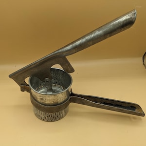 May include: A vintage, metal potato ricer with a long handle and a perforated bowl. The ricer is made of a silver-colored metal and has a sturdy, industrial design. The handle is angled for easy use, and the bowl is designed to hold potatoes for mashing.