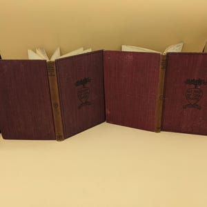 May include: Four antique books with burgundy covers and gold-toned spines are displayed open. The book covers feature a decorative emblem with the text "HEART OF OAK BOOKS". The pages are yellowed, suggesting age.