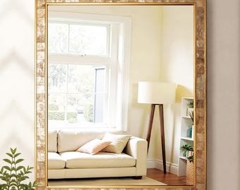 May include: A rectangular wall mirror featuring a wide frame composed of small, square, iridescent shell tiles in shades of beige and gold. The reflective glass surface displays a room interior with a white sofa, a floor lamp, and a window.