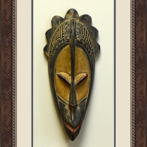 May include: A wooden mask with a dark brown and black finish. The mask has a stylized human face with a yellow painted face and a black painted nose and mouth. The mask has a decorative design around the top of the head.