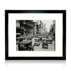 May include: Black framed print featuring a black and white photograph of a busy city street filled with vintage cars. The image captures a moment in time with buildings lining the street.