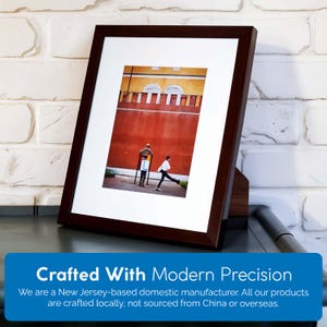 Pu&ograve; includere: Una fotografia incorniciata di un edificio in mattoni rossi con due figure che camminano di fronte ad esso. La foto &egrave; in una cornice marrone scuro con un passepartout bianco.