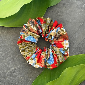 Può includere: Un colorato elastico per capelli con un motivo di ispirazione giapponese. Il tessuto presenta tonalità rosse, blu e oro con disegni floreali e geometrici. Questo accessorio per capelli è perfetto per aggiungere un tocco di colore a qualsiasi acconciatura.