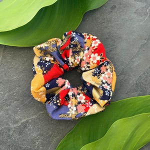 Può includere: Un colorato elastico per capelli con un motivo floreale. Il tessuto presenta un mix di tonalità rosse, blu navy, gialle e oro, con accenti di fiori rosa e bianchi. Accessorio per capelli.