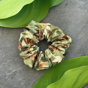 Può includere: Un elastico per capelli verde con un motivo floreale nei toni del marrone, oro e bianco. Il tessuto è arricciato ed elasticizzato, creando una forma circolare. Un accessorio per capelli.