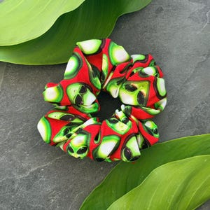 May include: A red scrunchie with a repeating pattern of green and white alien faces. The scrunchie is made of fabric and is designed to hold hair in place. The scrunchie is a fun and unique accessory.