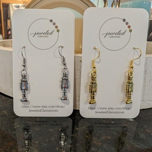 May include: Two pairs of nutcracker earrings on white card stock. The earrings are silver and gold. The card stock has the text "jeweled gemstones" and a website address.