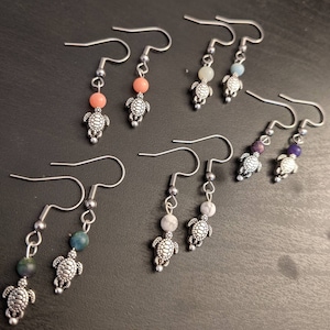 May include: A set of six pairs of silver turtle earrings with different colored gemstone beads. The beads are in shades of pink, blue, purple, and green.