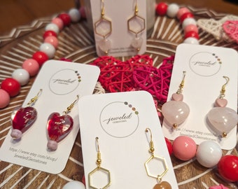May include: Four pairs of earrings displayed on white cards with the text "jeweled gemstones" on each card. The earrings are made of gold and feature different gemstone shapes, including hearts, hexagons, and a pink heart with a gold accent. The cards are placed on a wooden surface with pink and white beads and hearts.