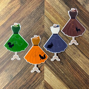 May include: Four paper doll cutouts of dresses with different colors and animal silhouettes. The dresses are green, orange, blue, and brown. The animals are a squirrel, a fox, a rabbit, and a deer.
