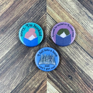 May include: Three blue and purple button pins with different book-themed illustrations and text. The first pin says "sorry, I'm booked" and features a cartoon woman reading a book. The second pin says "I could be social or I could read" and features a cartoon woman reading a book. The third pin says "I fell into a book and can't get out" and features a book with a forest and a house inside.