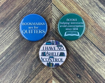 May include: Three round button pins with book-themed sayings. The blue pin says "BOOKMARKS are for QUITTERS". The green pin says "BOOKS helping introverts avoid conversation since 1454". The pink pin says "I HAVE NO SHELF CONTROL" with a bookshelf design.