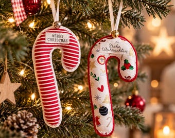May include: Two fabric candy cane ornaments hanging from white ribbons. The left ornament features red and white horizontal stripes with a tag reading Merry Christmas. The right ornament has a cream background with holiday icons and a tag reading Frohe Weihnachten.