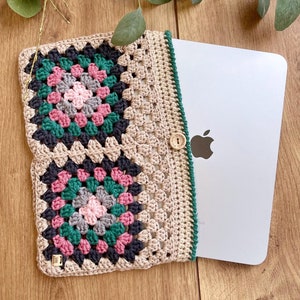 May include: A crocheted laptop sleeve with a granny square design in beige, black, green, and pink. The sleeve has a button closure and a green trim. The sleeve is shown with a laptop inside.