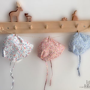 May include: Three floral baby bonnets in white, pink, and blue hang from a wooden rack. The bonnets are tied with strings and feature a delicate floral pattern. Wooden animal figurines decorate the top of the rack. The text "Floral & Dots" is visible.