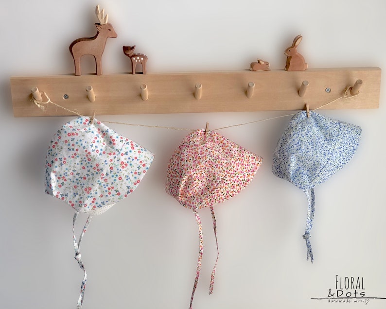 May include: Three floral baby bonnets in white, pink, and blue hang from a wooden rack. The bonnets are tied with strings and feature a delicate floral pattern. Wooden animal figurines decorate the top of the rack. The text "Floral & Dots" is visible.
