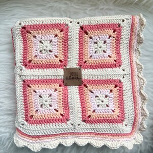 May include: A crocheted baby blanket in shades of pink, peach, and white with a scalloped edge. The blanket features a square pattern with a smaller square in the center of each. A brown leather tag with the text "Floral & Dots Handmade with Love" is attached to the blanket.