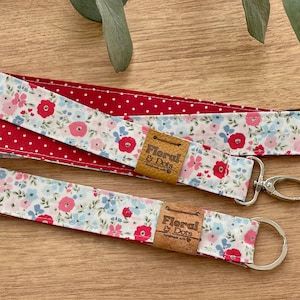 Könnte beinhalten: Zwei florale Schlüsselbänder und ein passender Schlüsselanhänger. Die Schlüsselbänder haben auf der einen Seite ein rotes Tupfenmuster und auf der anderen Seite einen weißen Hintergrund mit rosa und blauen Blumen. Jedes hat ein Lederetikett mit der Aufschrift "Floral & Dots."