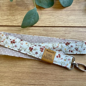 Può includere: Un portachiavi in tessuto bianco e beige con un motivo floreale e un anello portachiavi argentato. Il portachiavi ha un'etichetta in pelle marrone con il testo "Floral & Dots" su di esso.