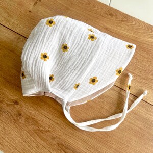 May include: White baby bonnet with yellow sunflower embroidery. The bonnet is made of a textured fabric and features a tie closure. The bonnet is designed to protect the baby's head from the sun and wind.