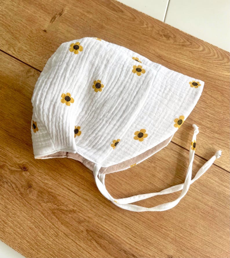 May include: White baby bonnet with yellow sunflower embroidery. The bonnet is made of a textured fabric and features a tie closure. The bonnet is designed to protect the baby's head from the sun and wind.