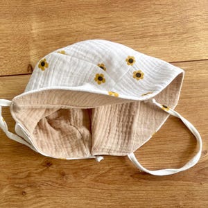 May include: White baby sun hat with a yellow sunflower pattern. The hat has a light tan interior and white ties. The hat is made of a soft, textured fabric. The hat is designed to protect the baby's face from the sun.