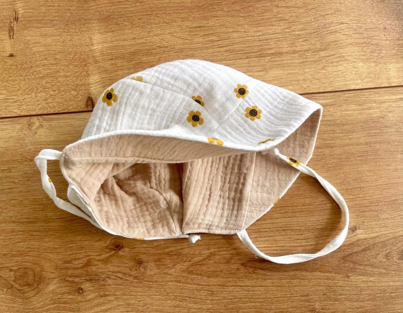 May include: White baby sun hat with a yellow sunflower pattern. The hat has a light tan interior and white ties. The hat is made of a soft, textured fabric. The hat is designed to protect the baby's face from the sun.