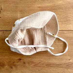 May include: A white and beige baby sun hat with a sunflower pattern. The hat is made of soft, textured fabric and features a drawstring. The interior is a solid beige color. The hat is designed to provide sun protection.