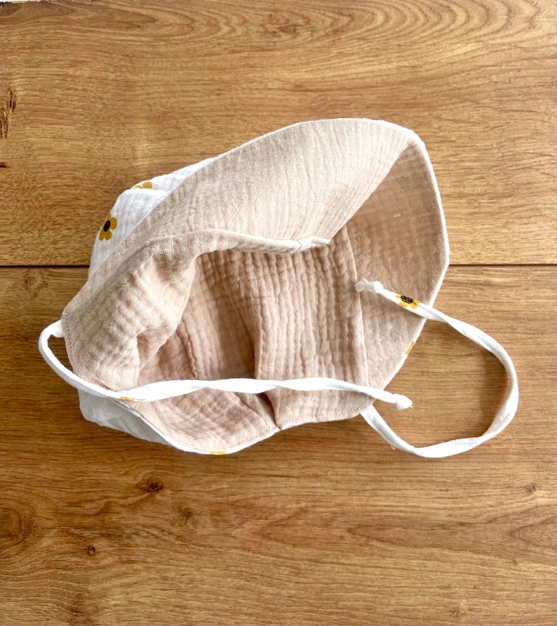 May include: A white and beige baby sun hat with a sunflower pattern. The hat is made of soft, textured fabric and features a drawstring. The interior is a solid beige color. The hat is designed to provide sun protection.