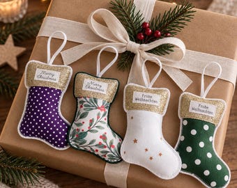 May include: Four small fabric Christmas stocking ornaments with white hanging loops. Two feature polka dots in purple and green, one has a botanical holly print, and one is white with gold stars. Each has a gold glitter cuff with printed text.