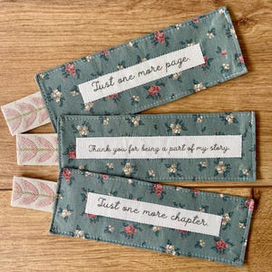 Könnte beinhalten: Drei Stoff-Lesezeichen mit floralen Mustern und weißen Bandakzenten. Jedes Lesezeichen hat ein weißes rechteckiges Etikett mit dem Text: "Just one more page", "Thank you for being a part of my story" und "Just one more chapter."
