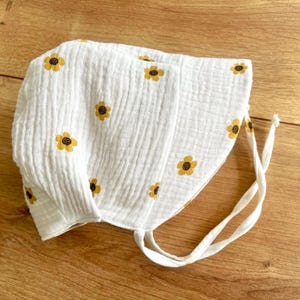 May include: White baby bonnet with yellow sunflower embroidery. The bonnet is made of a crinkled fabric and has a tie closure. The bonnet is designed to protect the baby's head from the sun.
