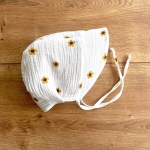 May include: White baby bonnet with yellow and brown floral pattern. The bonnet is made of a textured fabric and has white ties. The bonnet is designed to protect the baby's head from the sun.
