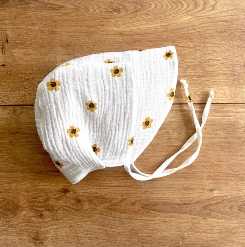 May include: White baby bonnet with yellow and brown floral pattern. The bonnet is made of a textured fabric and has white ties. The bonnet is designed to protect the baby's head from the sun.